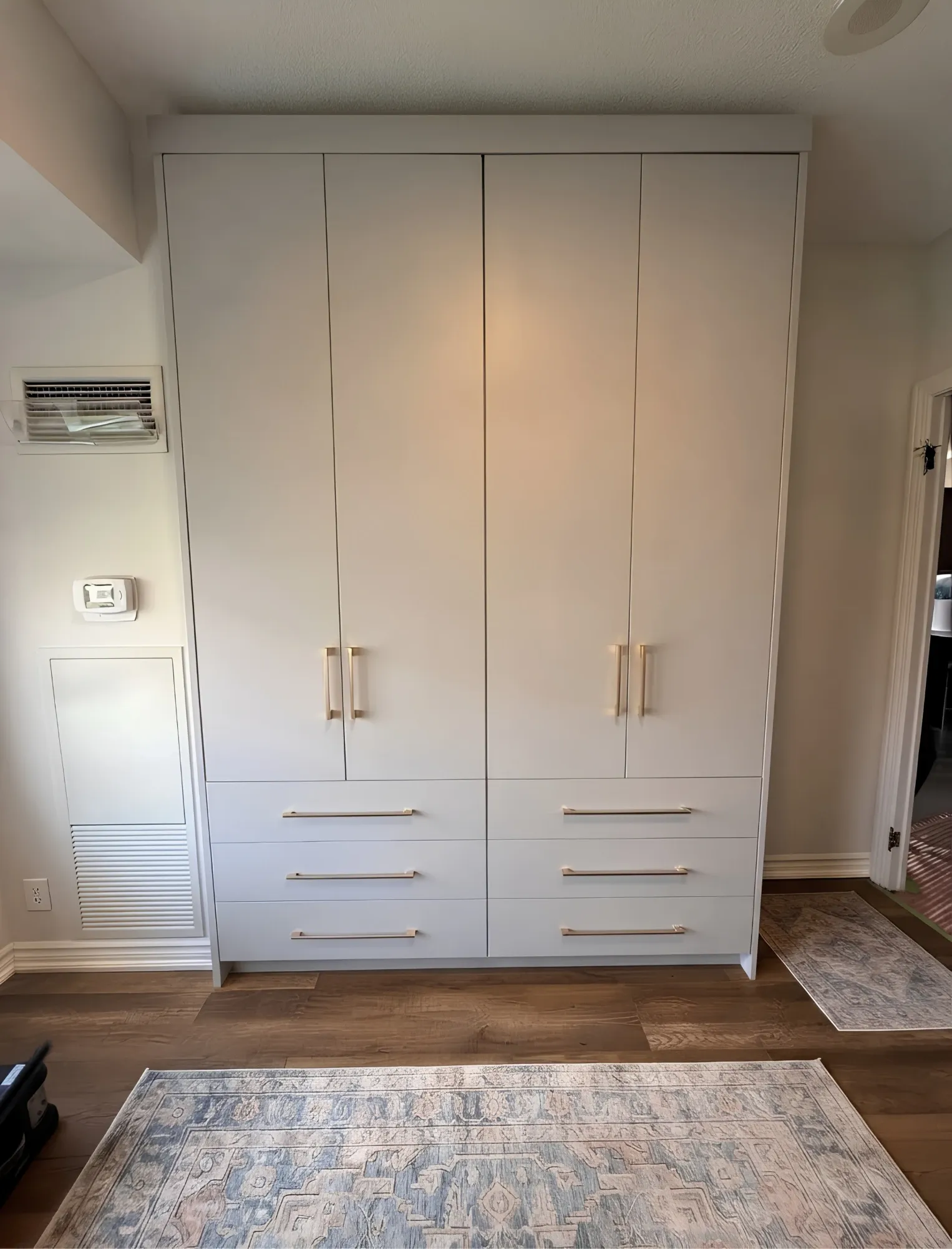 A four-door white wardrobe built to the ceiling beside a bedroom doorway, six drawers below and long brass pulls throughout