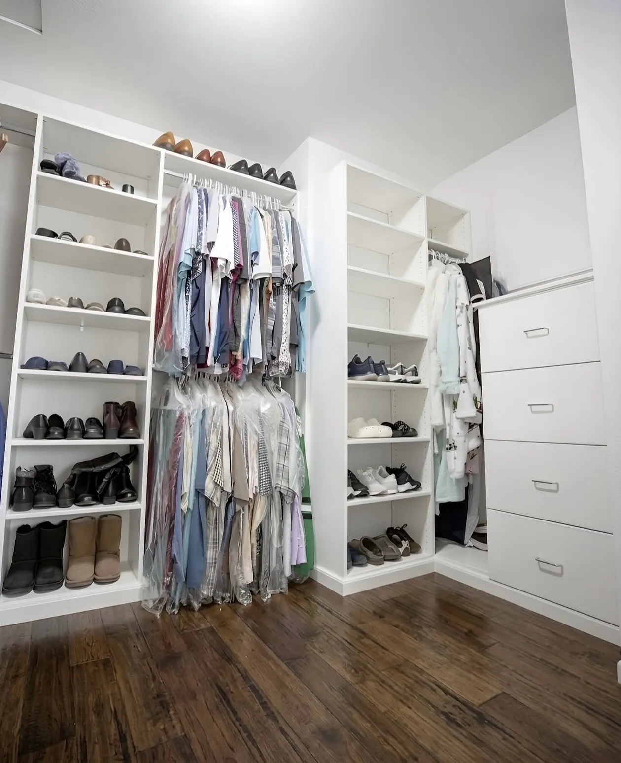 A white corner walk-in closet with clothes hung in garment bags at two levels, shoe shelving to the ceiling and a bank of drawers