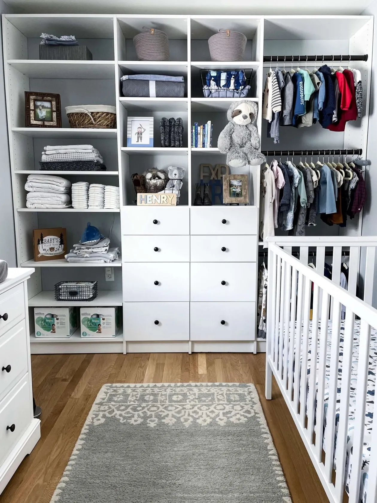 A nursery closet with two rails of baby clothes, eight drawers, and open shelving for books, blankets and toys beside the crib