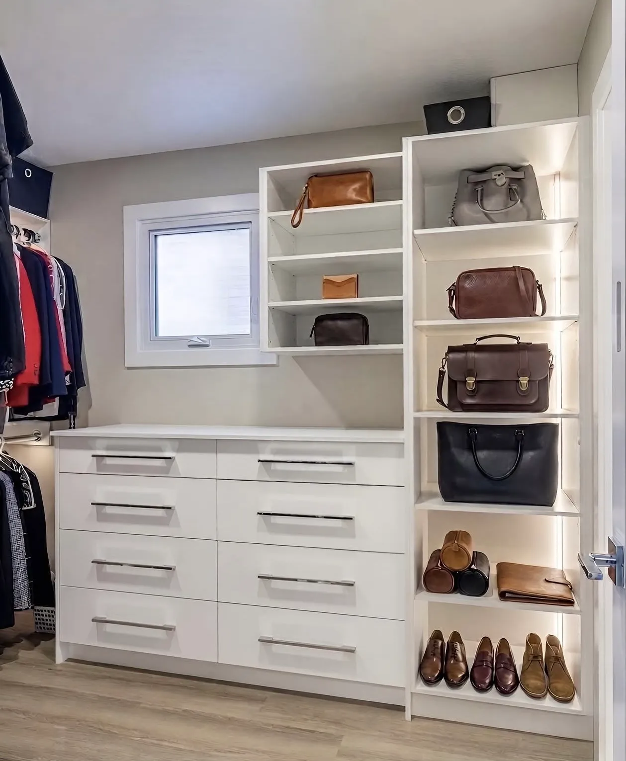 A white built-in wardrobe with a bank of eight drawers and a lit display tower holding handbags and shoes