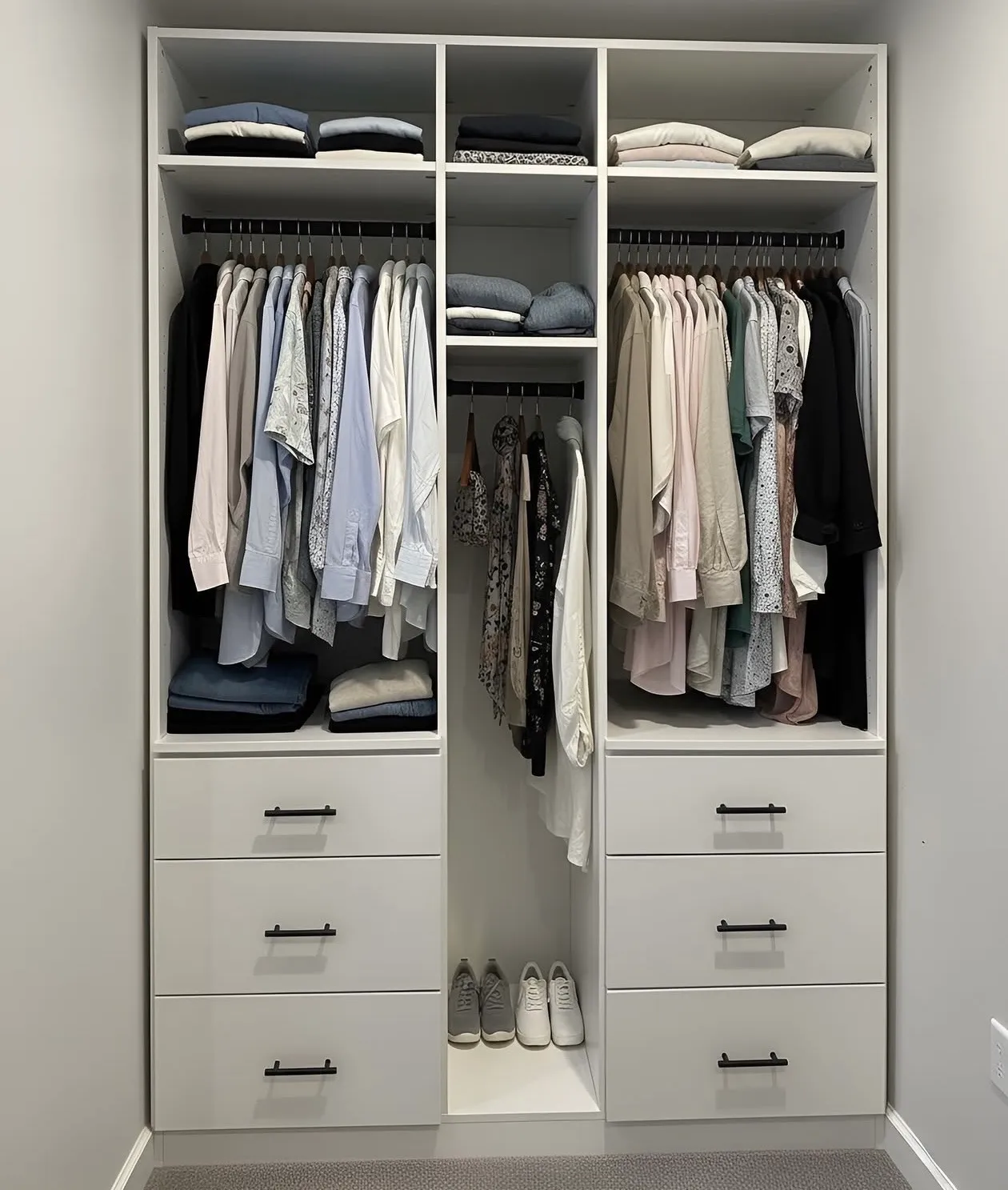 A white reach-in closet with two hanging sections either side of a centre tower, over two banks of drawers with black bar pulls