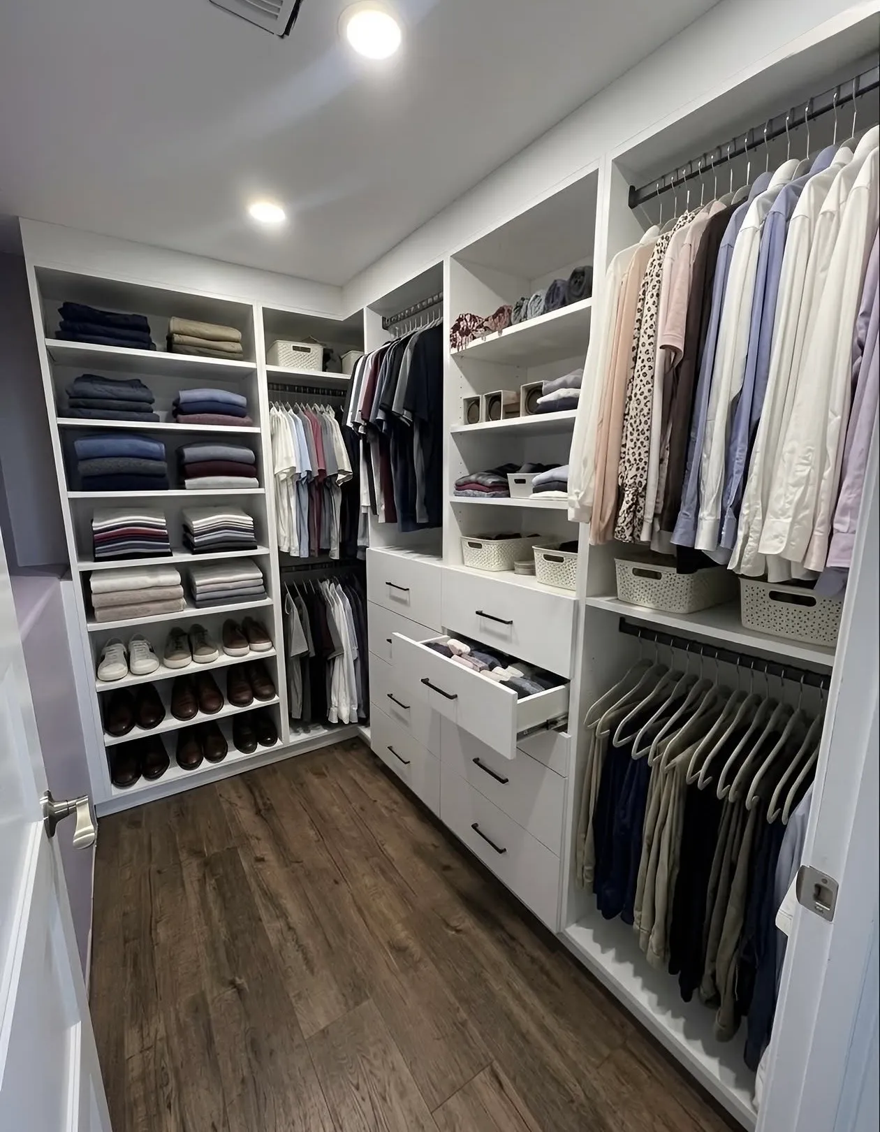 A white galley walk-in closet with folded shelving down one side, a centre drawer bank pulled open and hanging on both walls