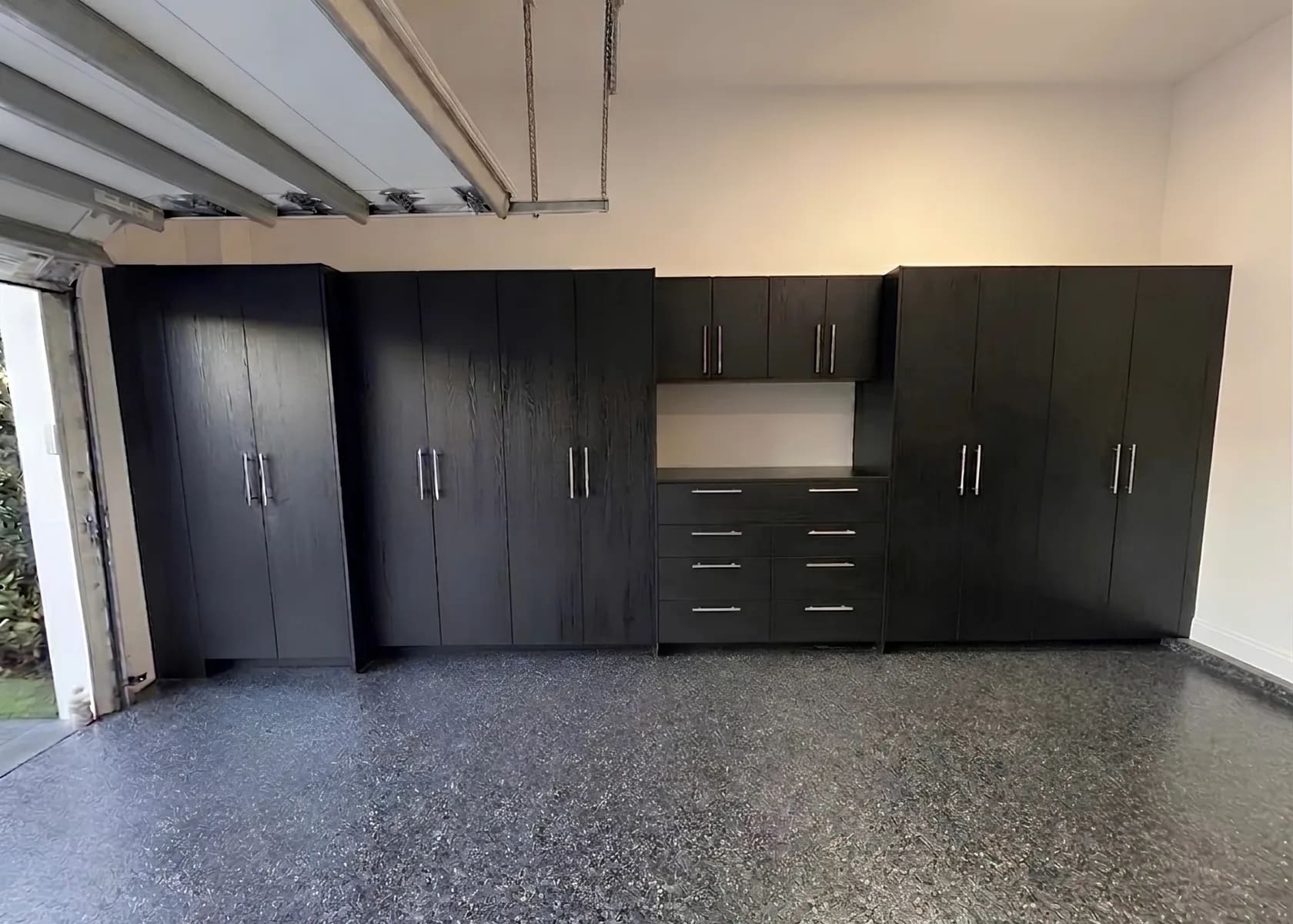Black garage cabinets across the back wall of a garage — full-height units either side of a central drawer bank with a worktop and upper cabinets above it