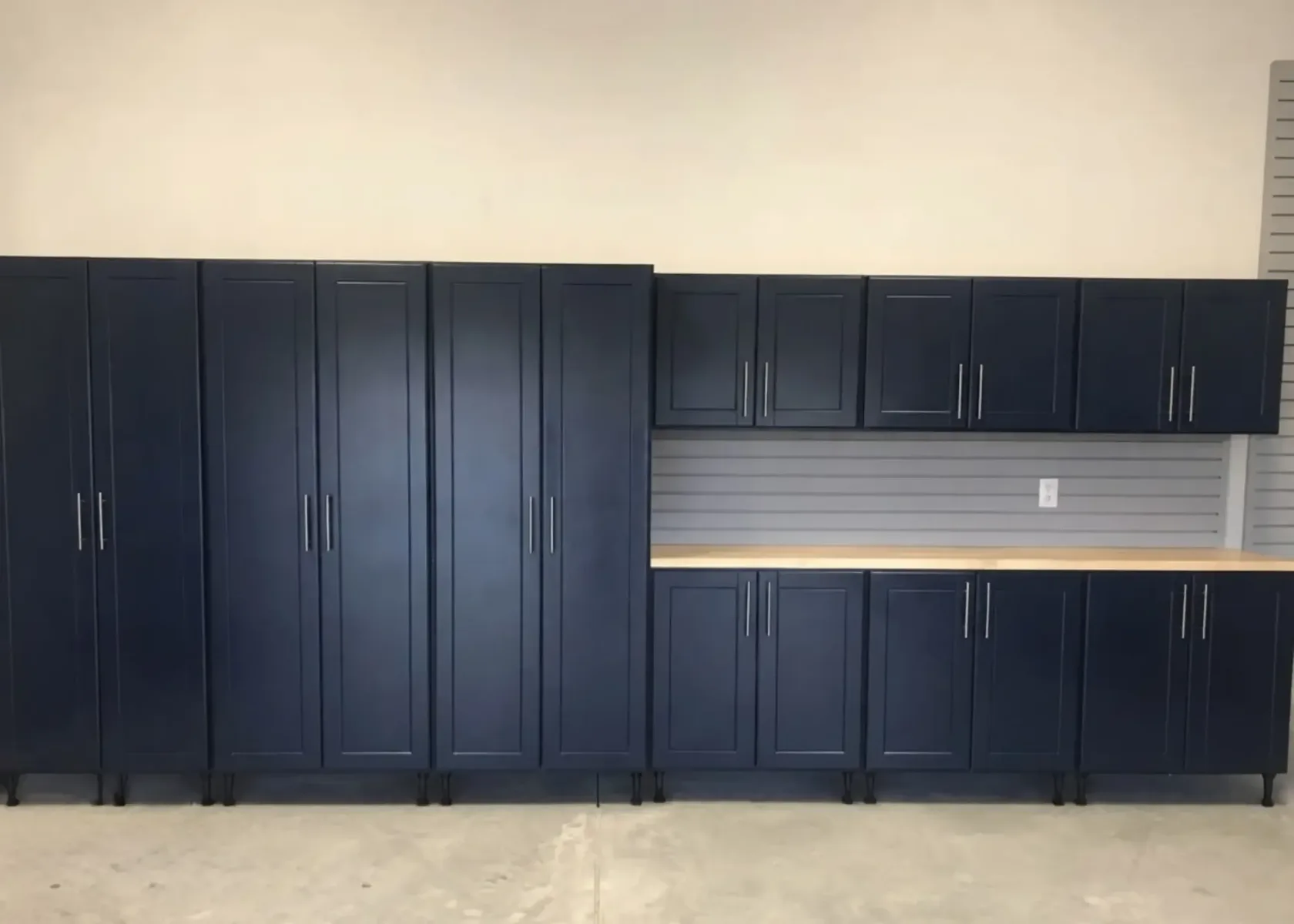 Navy blue garage cabinets on adjustable levelling legs, with grey slatwall panelling between the upper cabinets and a butcher-block worktop