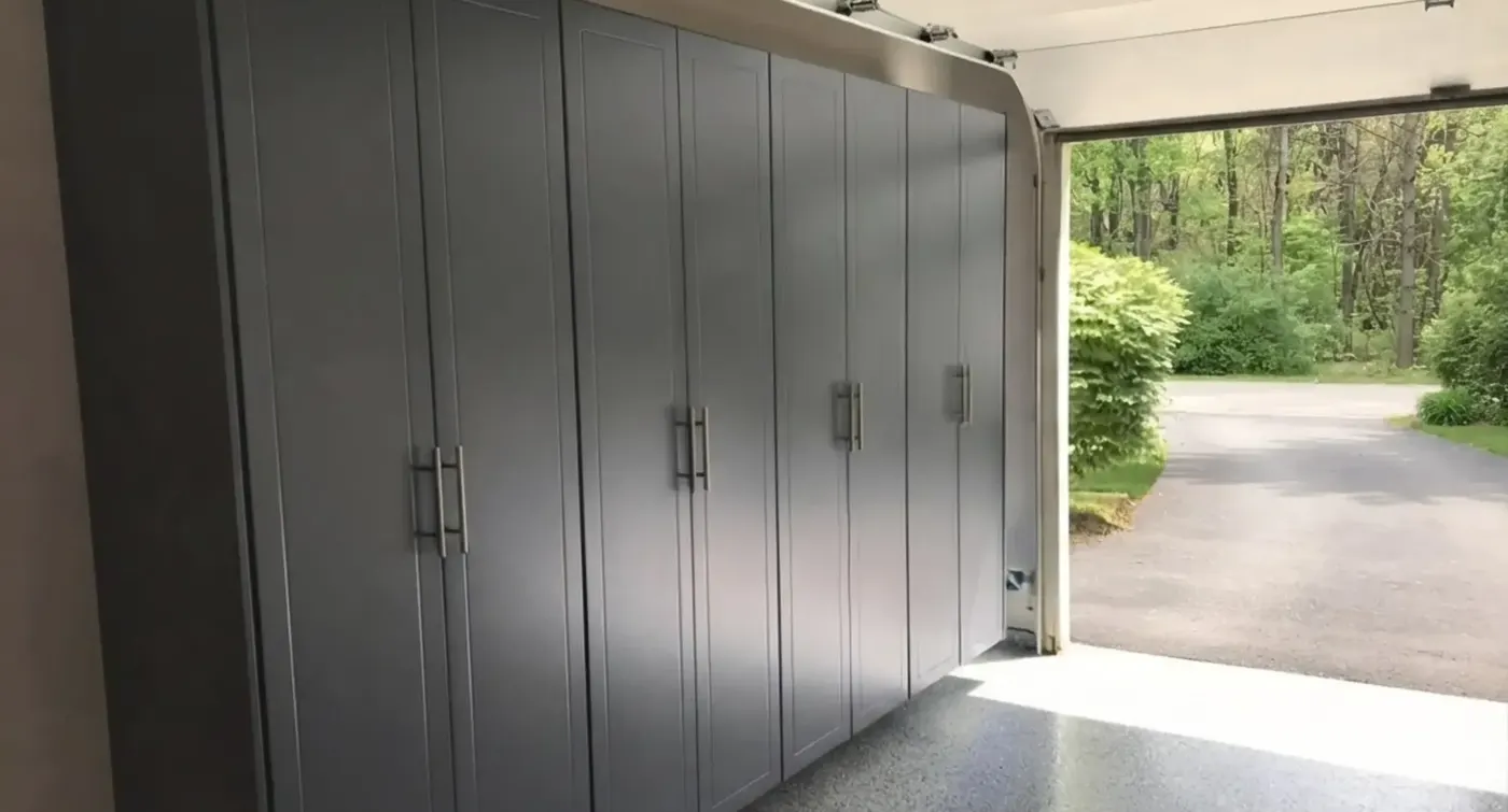 Grey shaker garage cabinets running the full length of a garage wall, with the garage door open to a driveway and trees beyond