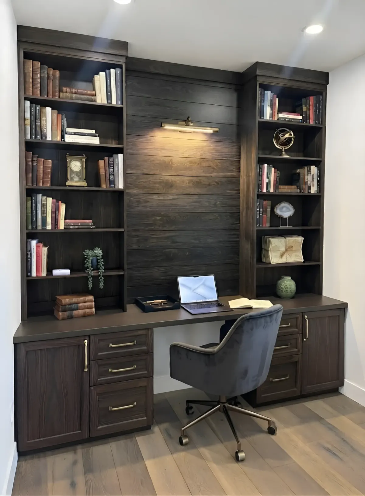 A dark espresso home office - two full-height bookcases either side of a shiplap back panel under a brass picture light, a desk across the wall and drawers and cupboards beneath it
