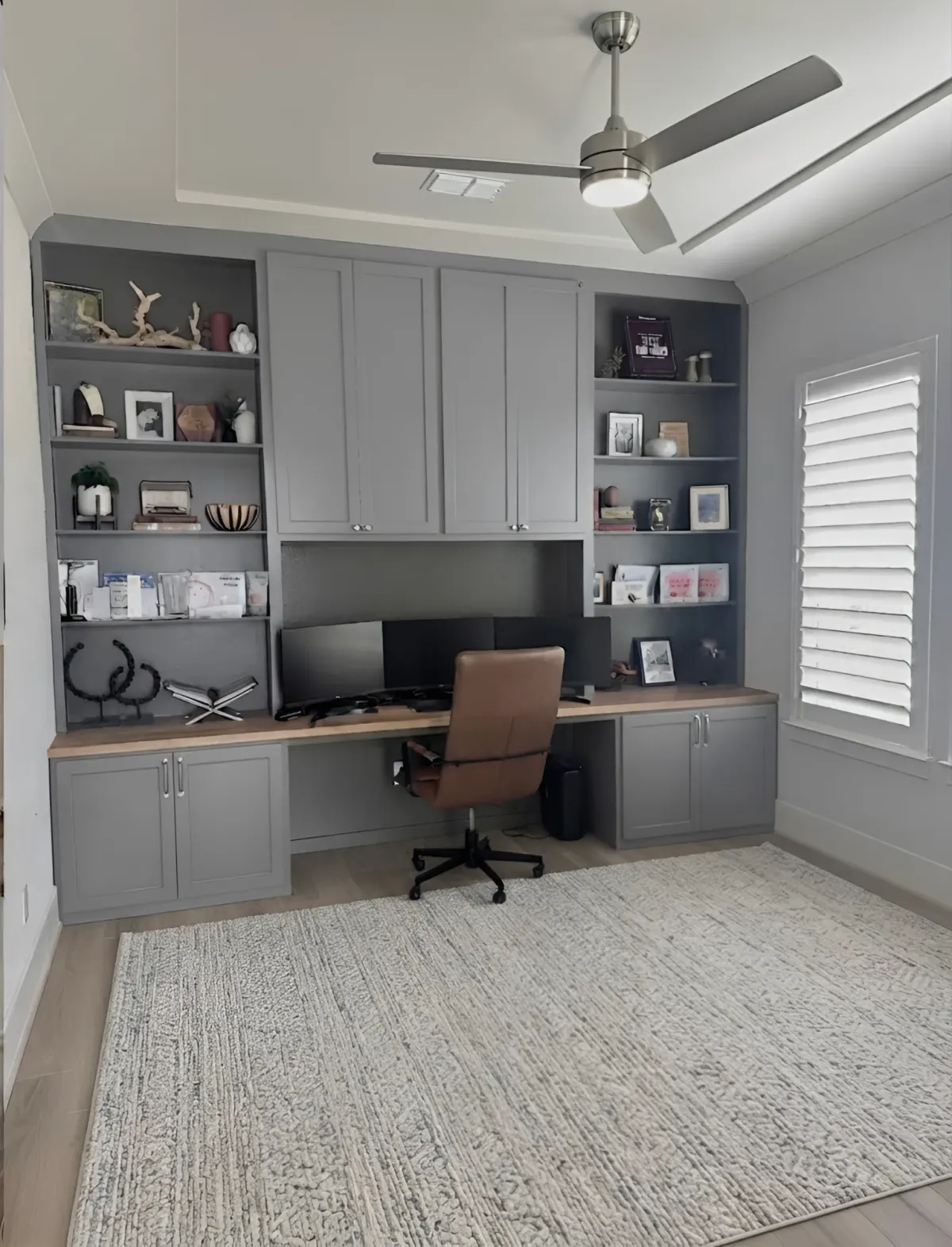 A grey painted home office wall - closed uppers over the desk, open display shelving either side and a timber work surface carrying two monitors over cabinets