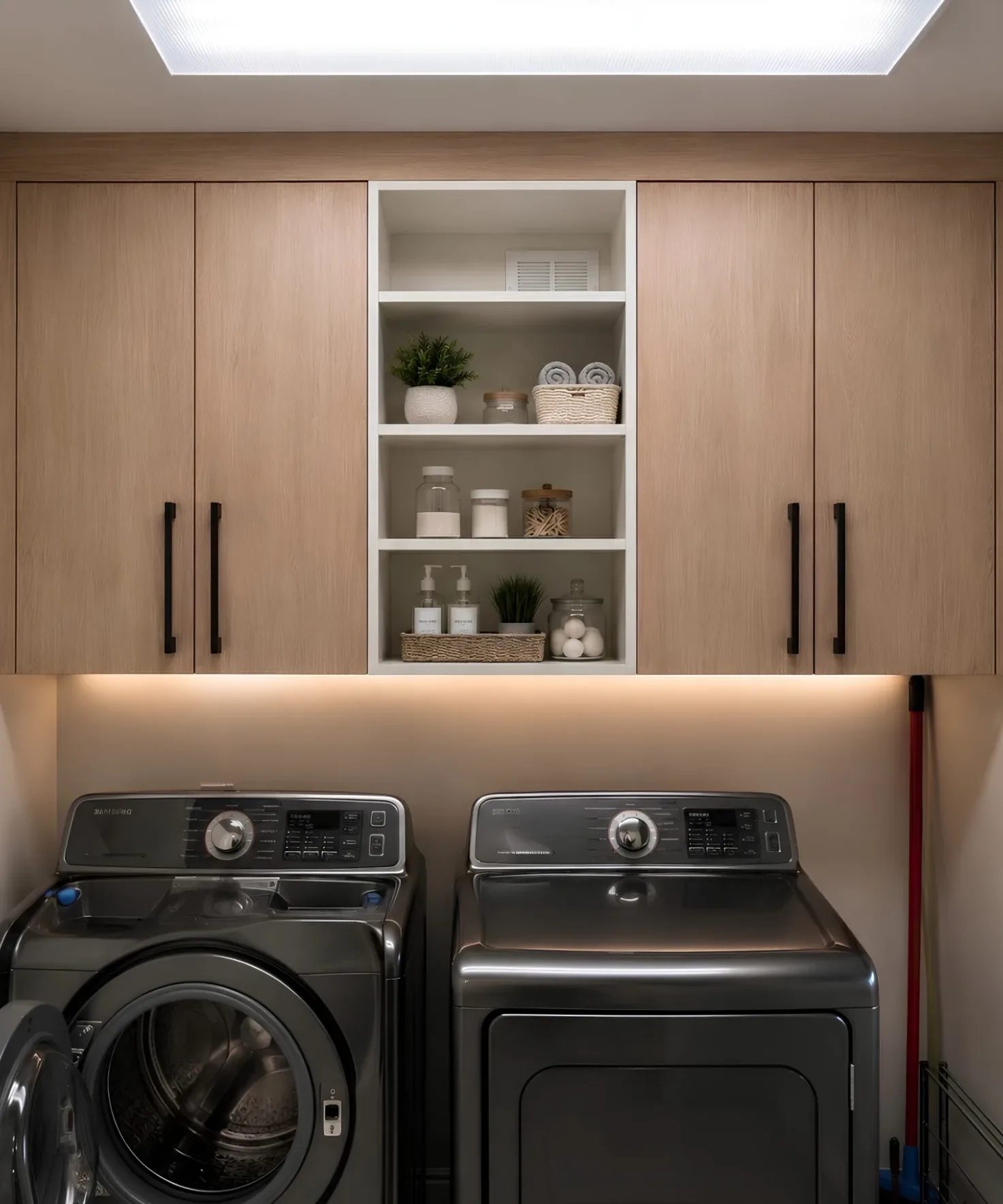 Pale oak wall cabinets either side of a white open shelving bay holding jars and folded towels, LED lighting under the run and a top-loading washer and dryer beneath