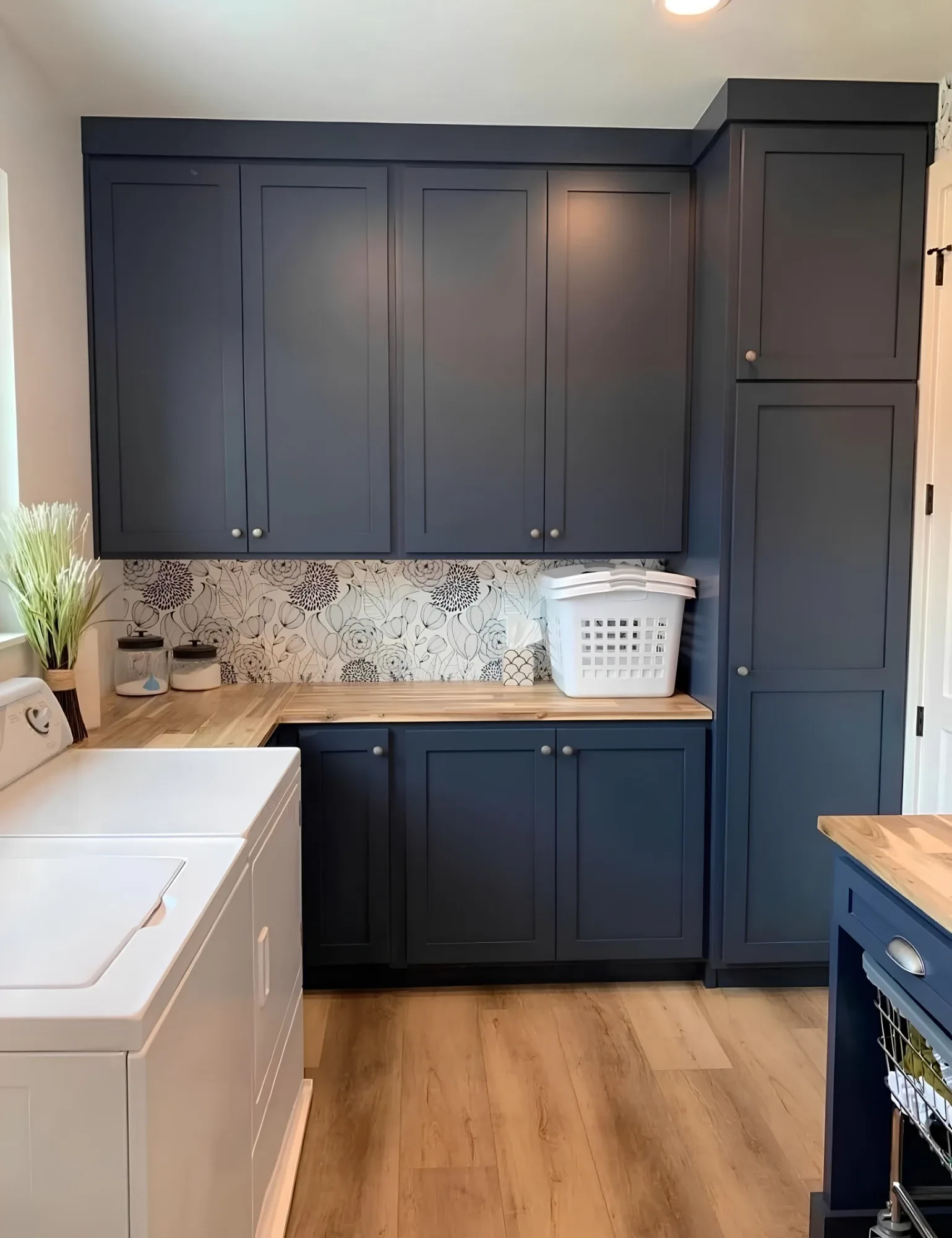 Navy shaker cabinets above and below a butcher block counter that turns a corner, a floral backsplash behind it and a full-height cupboard at the end of the run