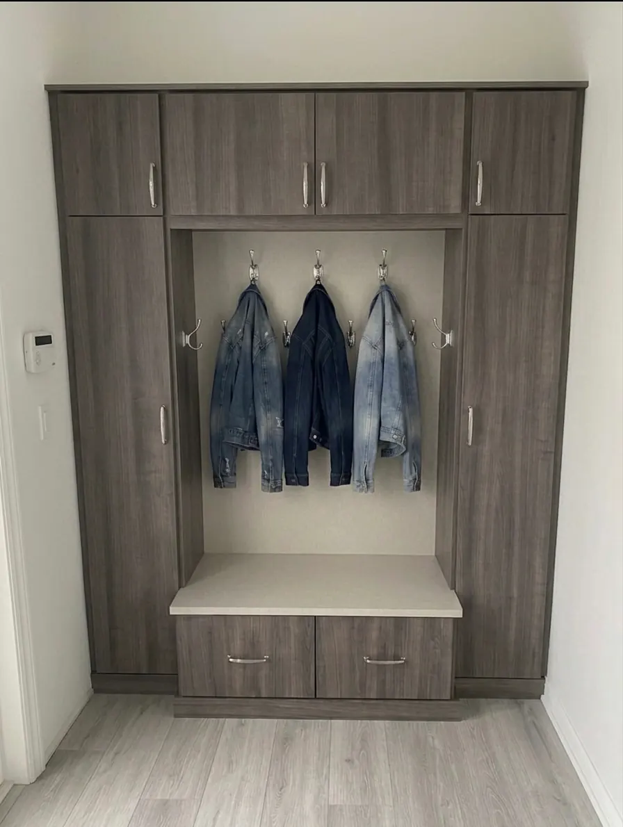 A grey woodgrain entryway unit with hooks and three denim jackets in the centre bay, full-height cupboards either side and a bench over two drawers