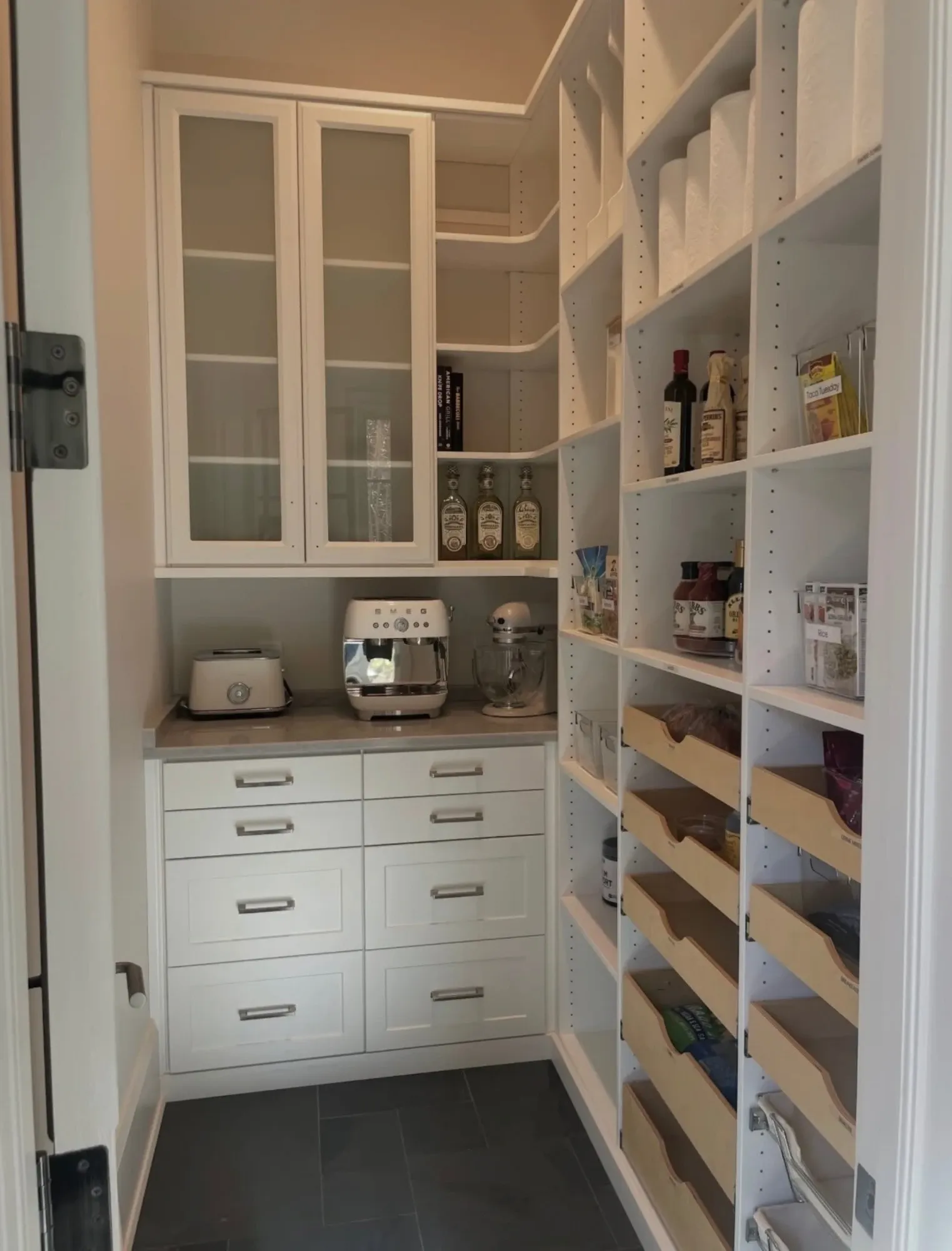 A cream pantry with glass-fronted upper cabinets over a counter holding a toaster, espresso machine and stand mixer, deep drawers beneath and a wall of open shelving with timber pull-out trays