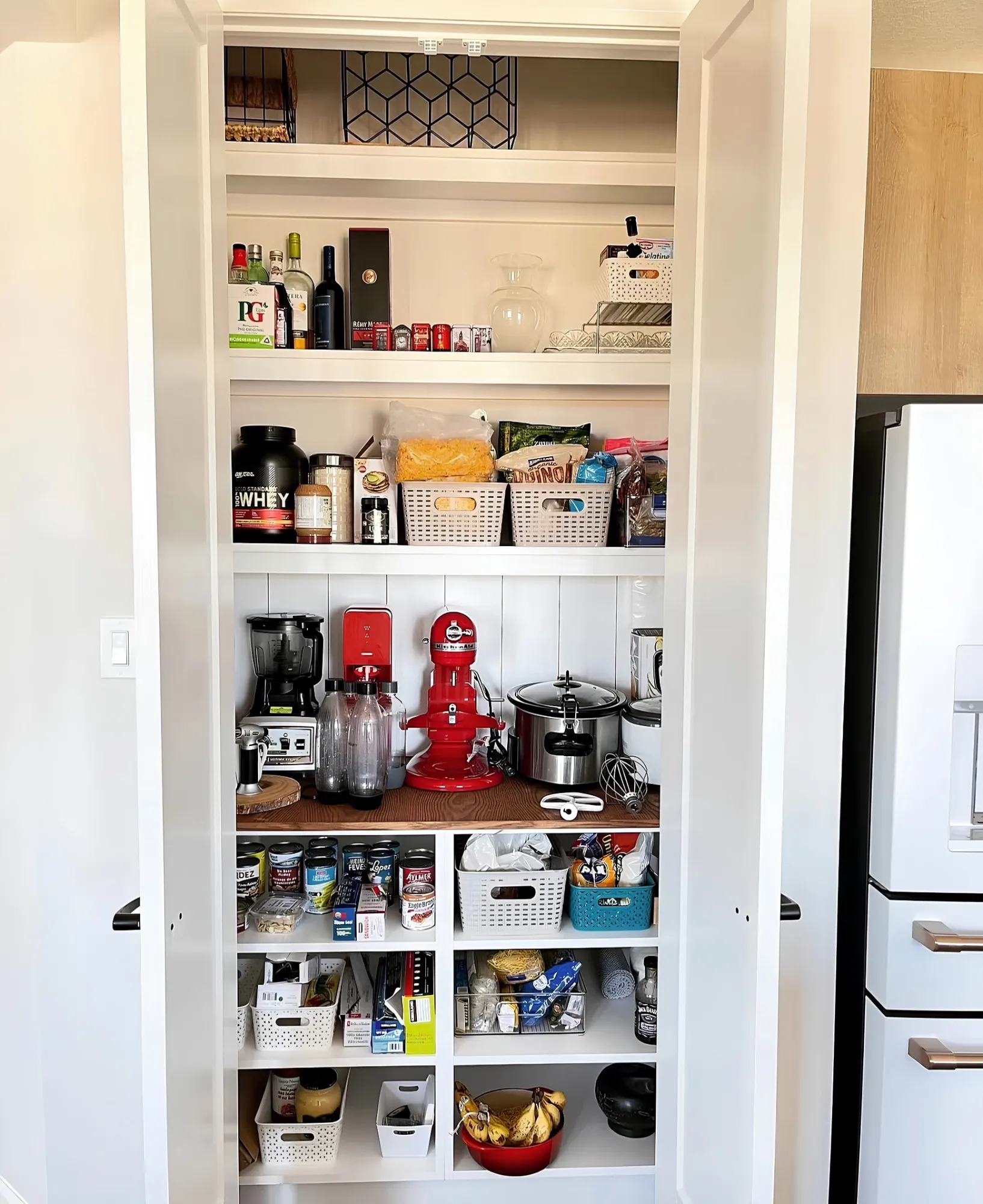 A white reach-in pantry with a timber counter for the mixer, blender and slow cooker, tins and baskets on the shelves above and below