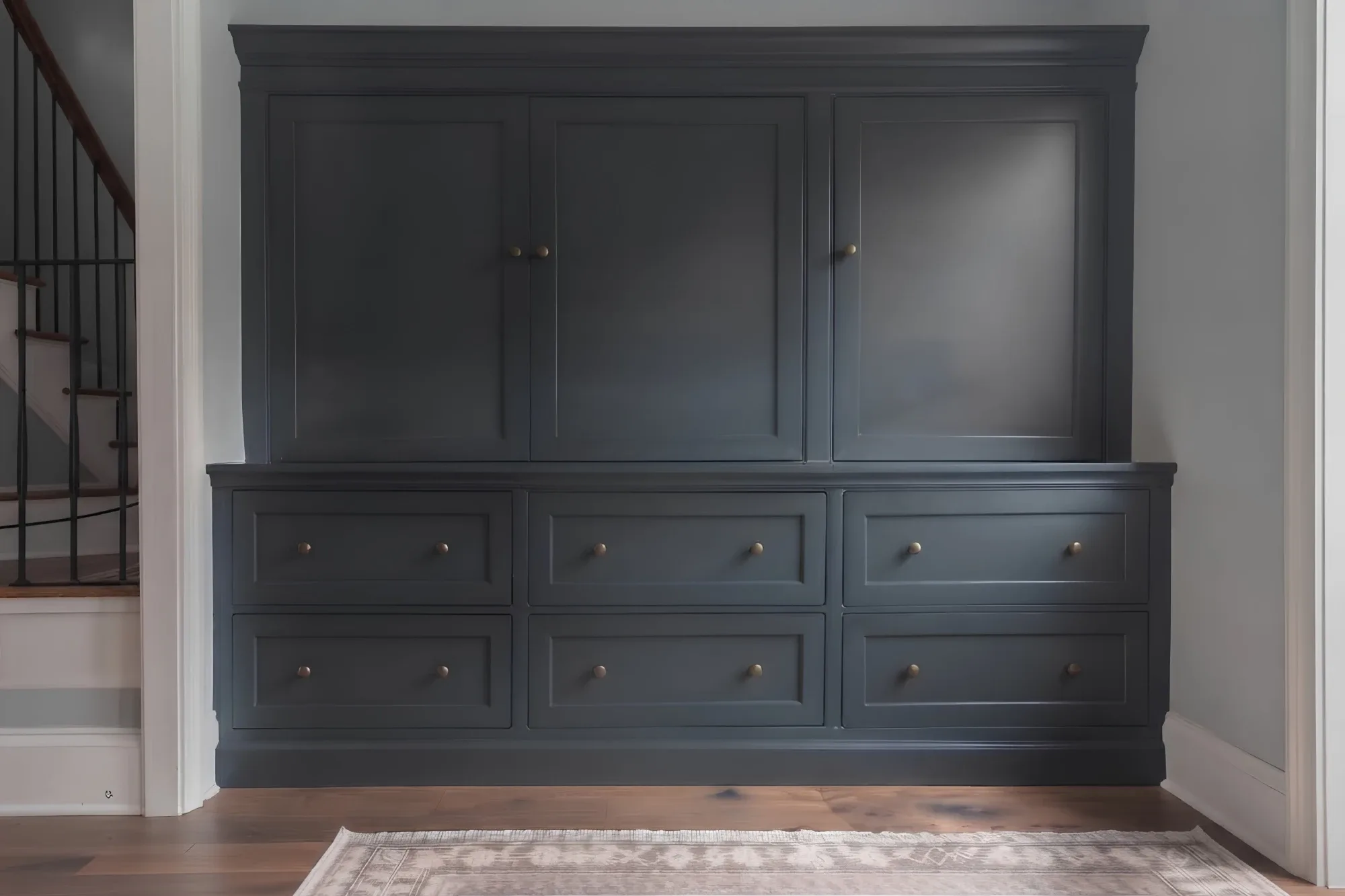 A charcoal built-in wall of cabinetry: three panelled upper doors above six panelled drawers with brass knobs, crown moulding at the top and base moulding to the floor