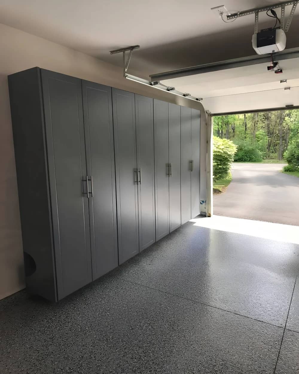 A full-height run of grey garage cabinets down one wall of a garage, floor to ceiling, on a speckled epoxy floor, with the door open to a driveway and trees beyond