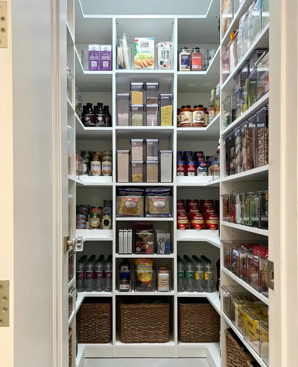 A white walk-in pantry shelved on three walls, with clear stacking bins and canisters at eye level, tinned goods and bottles below and woven baskets on the floor shelf
