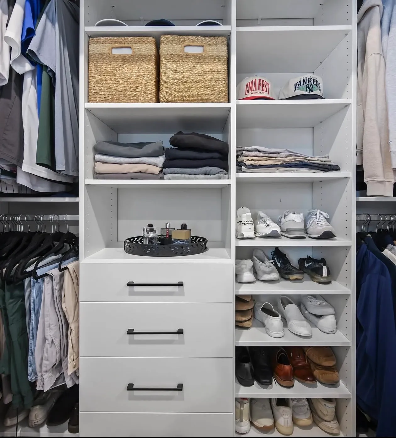 A white reach-in closet with hanging either side of a centre tower, baskets and folded shelving above three drawers and shoes on adjustable shelves