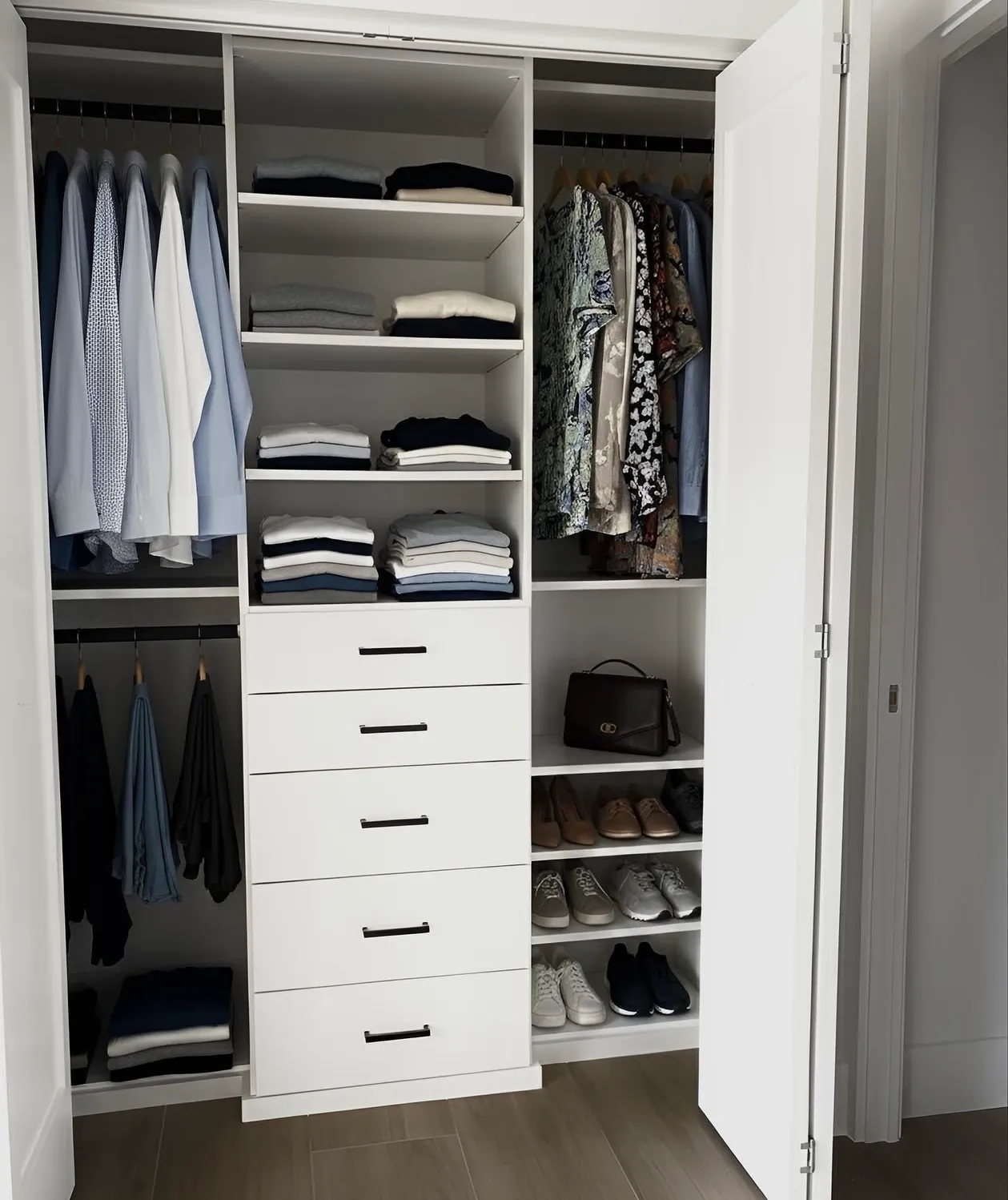 A white reach-in closet behind bifold doors, double hanging either side of a five-drawer tower under folded shelving, with shoes to the right