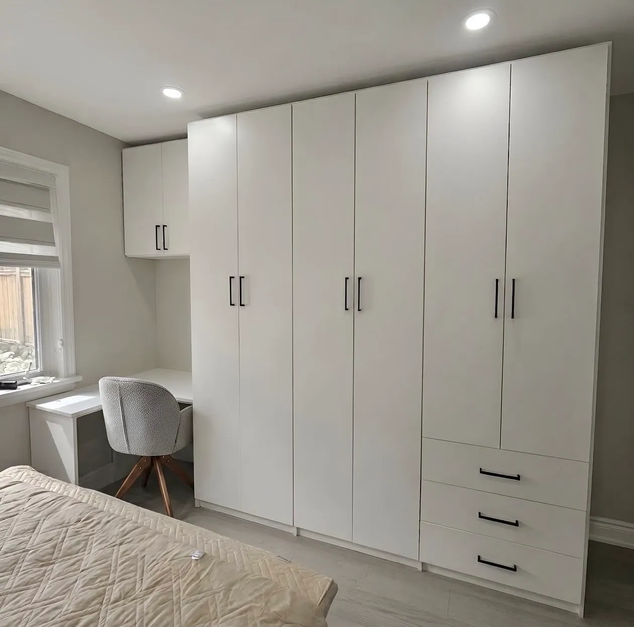 A white wardrobe wall run to the ceiling along one side of an Etobicoke bedroom, with a desk and cabinets built into the corner beside the window