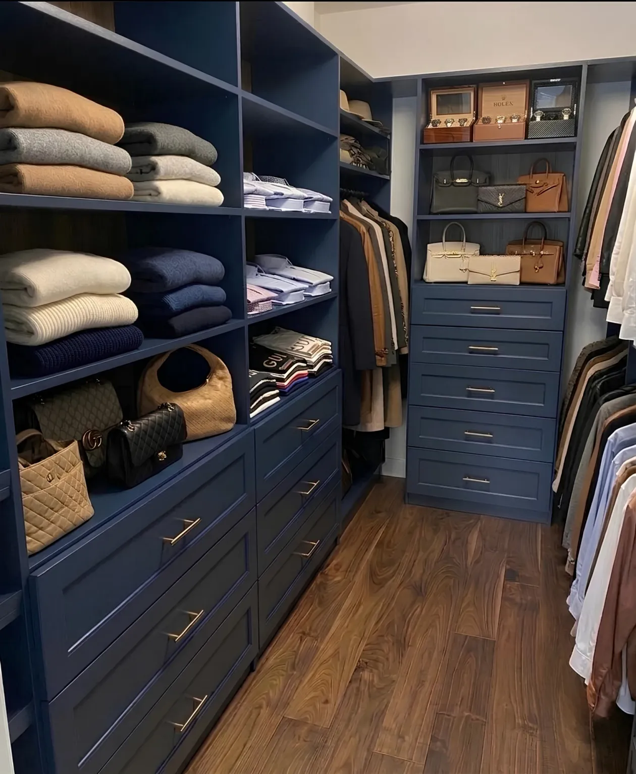 A navy walk-in closet with brass pulls, open shelving over banks of drawers, handbags displayed on the shelves and hanging down both walls