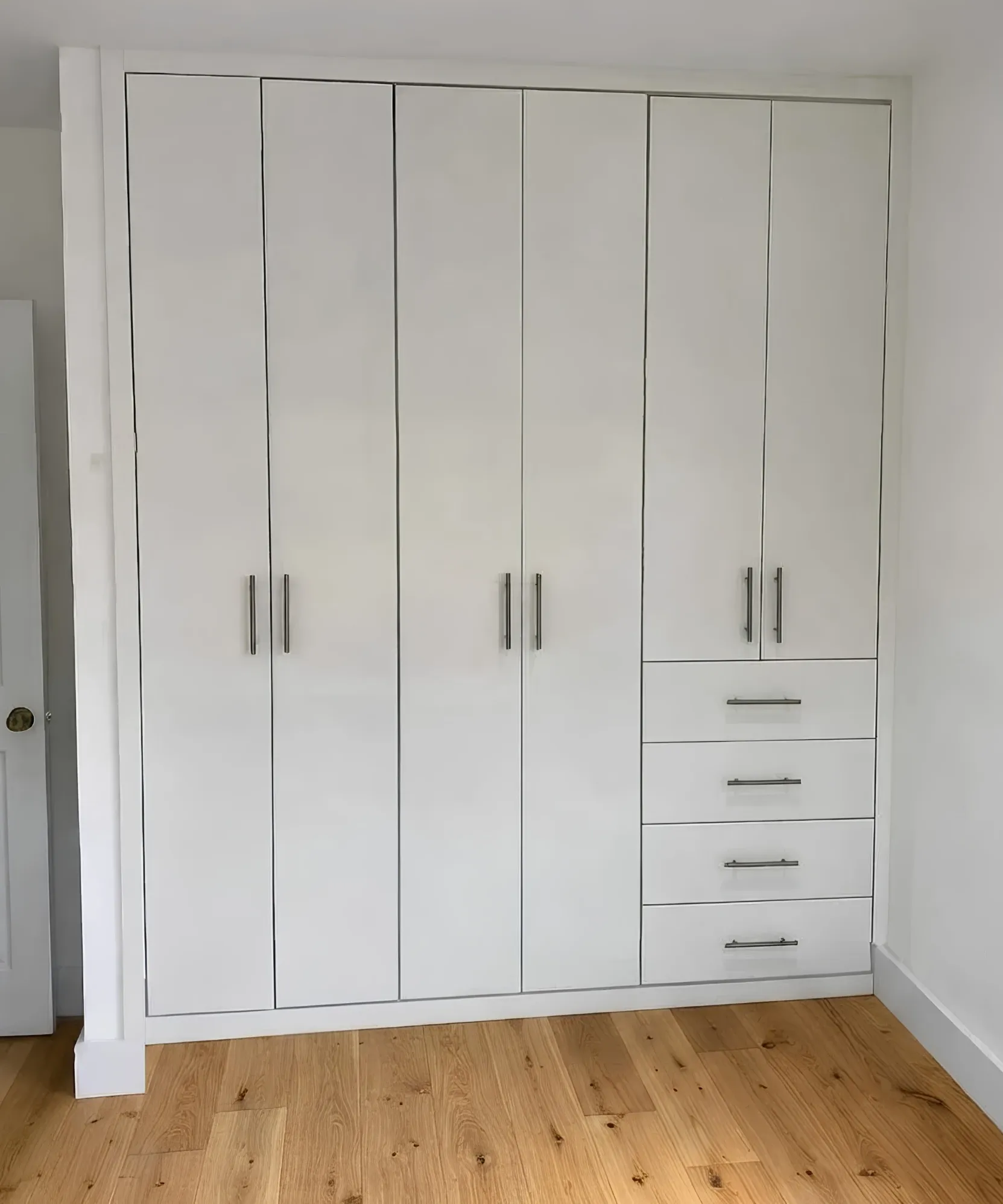 A white wardrobe built floor to ceiling into a bedroom corner, four full-height doors beside a bank of four drawers under a cupboard