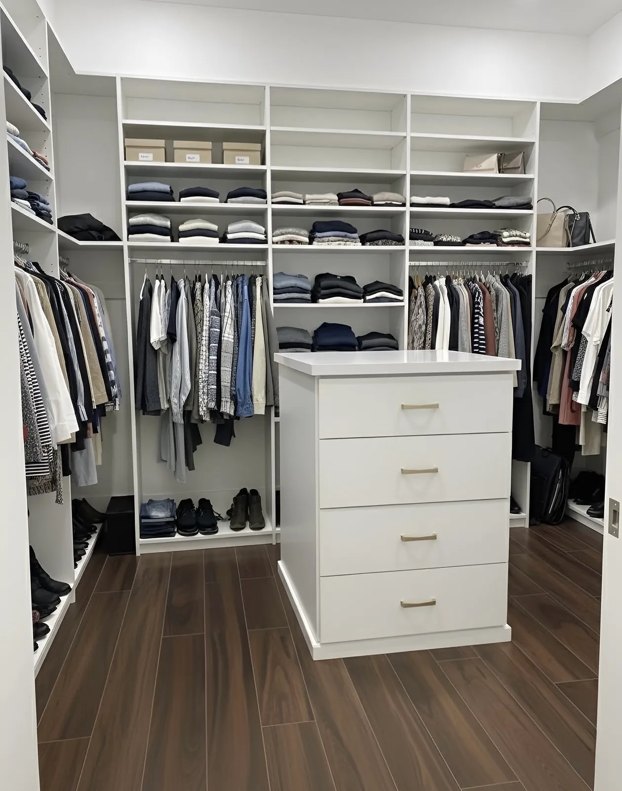 A white walk-in closet built out on all four walls with hanging at two levels under folded shelving, and a four-drawer island standing in the middle