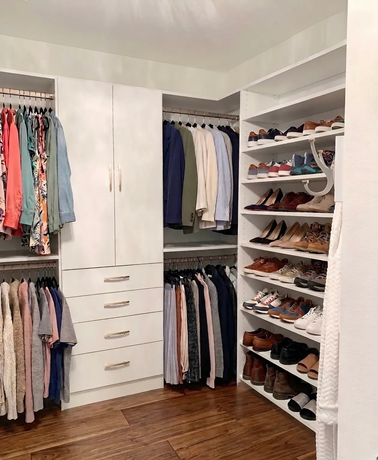 A corner walk-in closet with double hanging on two walls, a drawer bank with a cabinet over it, and a full shoe wall