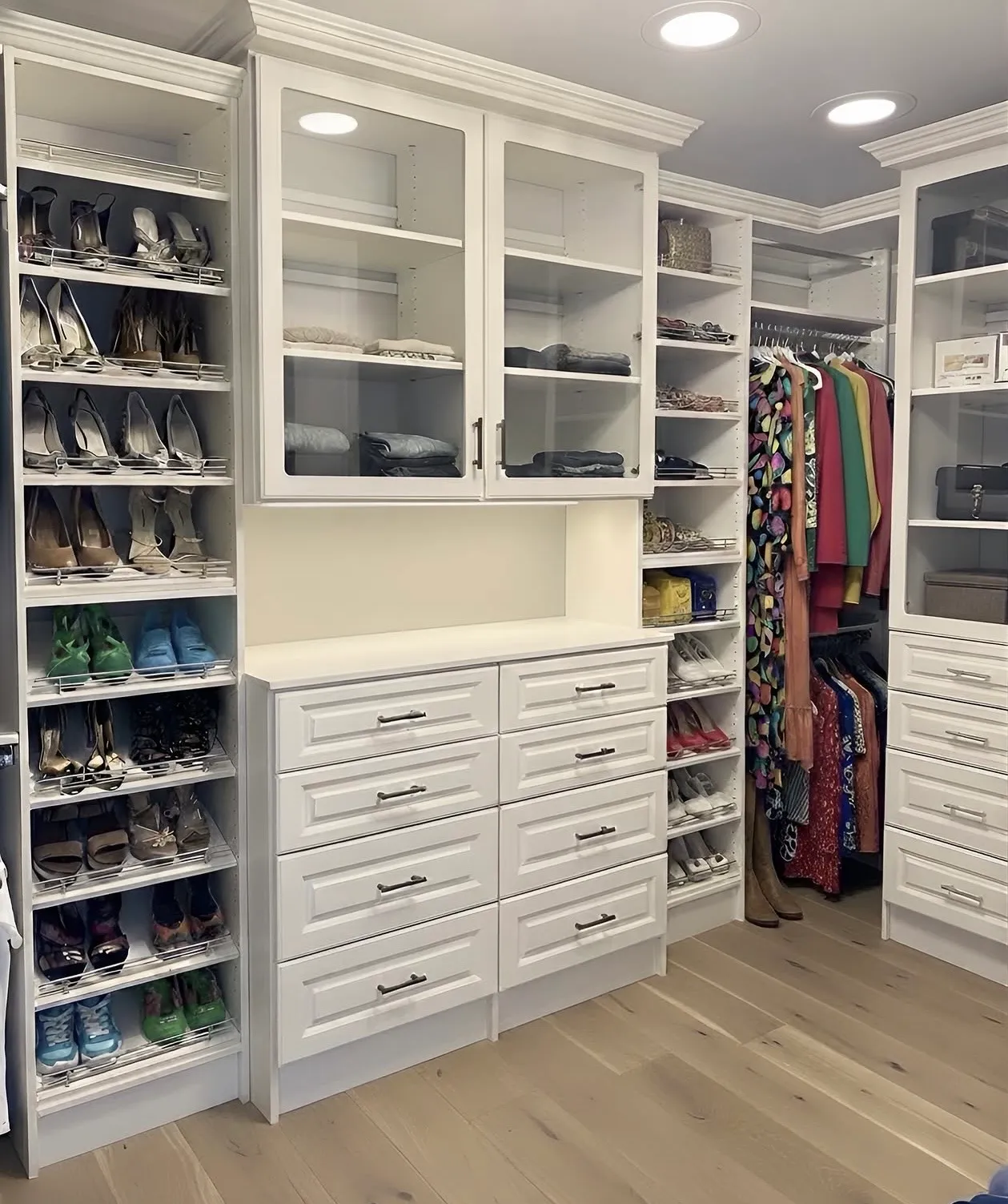 A large walk-in closet with glass-fronted upper cabinets over a drawer bank, pull-out shoe racks and hanging beyond