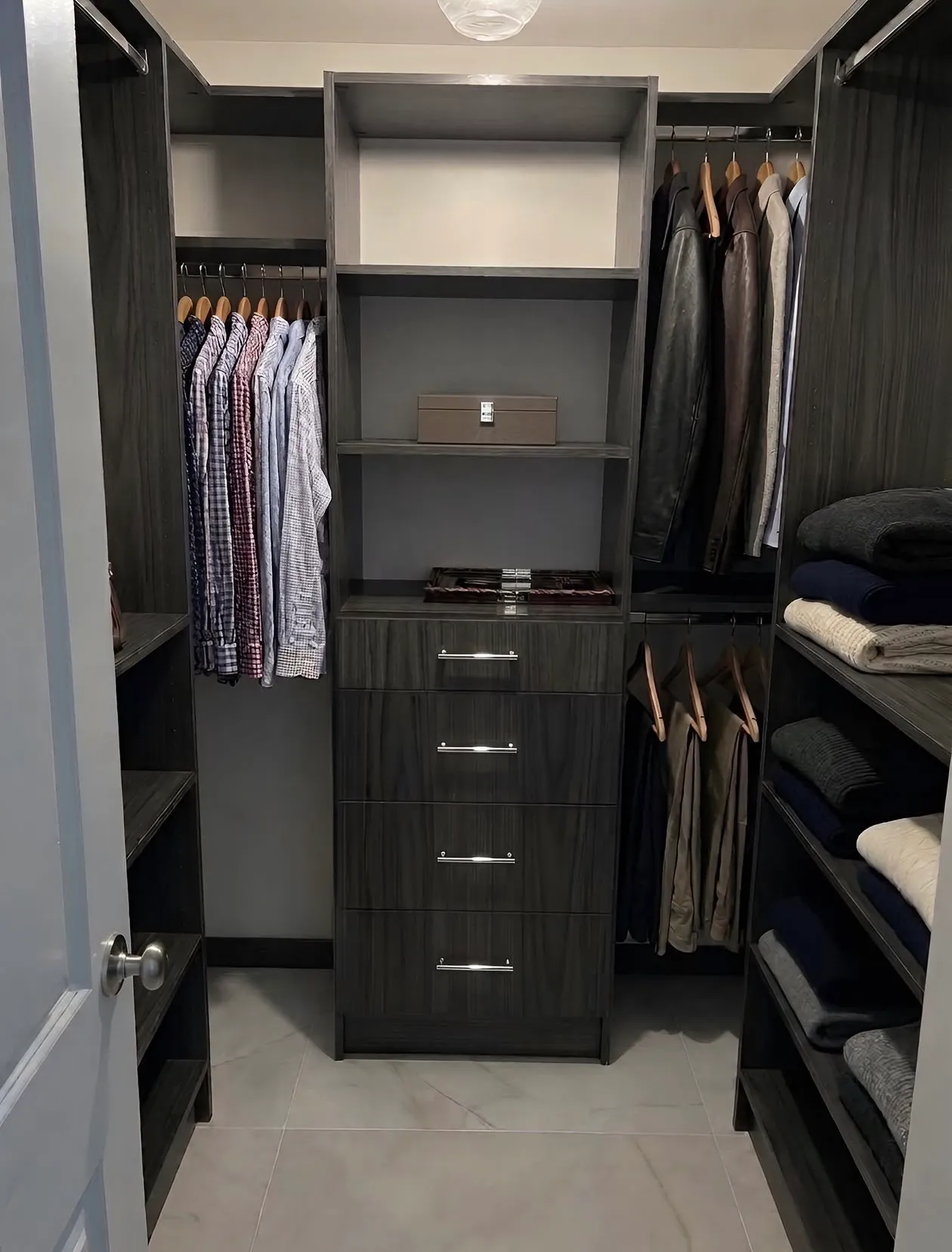 A compact dark oak walk-in closet with hanging on three walls either side of a four-drawer tower under open shelving