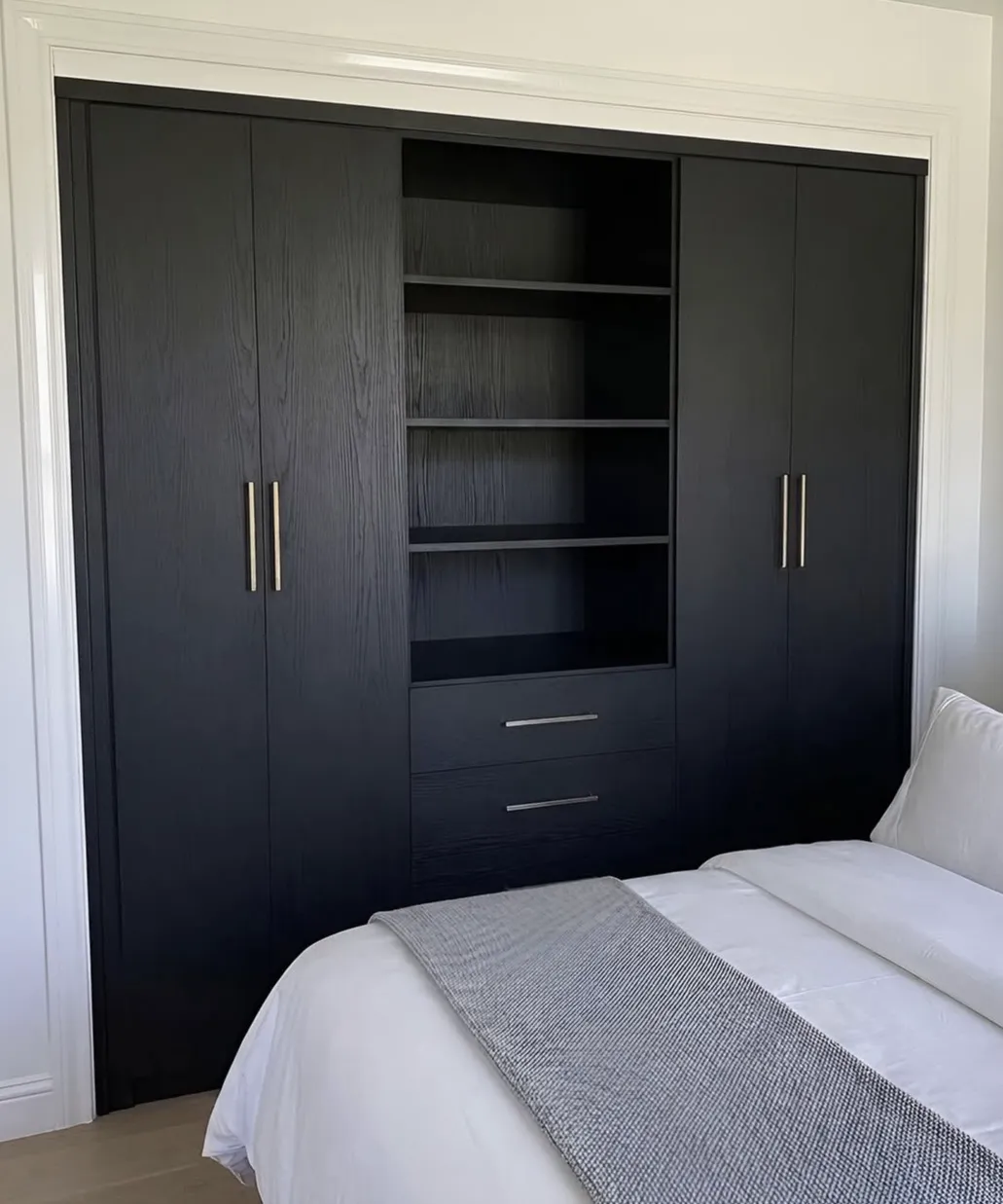 A black oak wardrobe wall built into a condo bedroom alcove, full-height doors either side of open shelving over two drawers