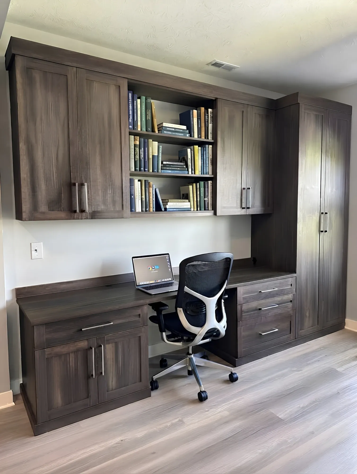 A dark walnut home office wall: upper cabinets either side of open bookshelves, a desk run between them and a drawer bank beside the chair