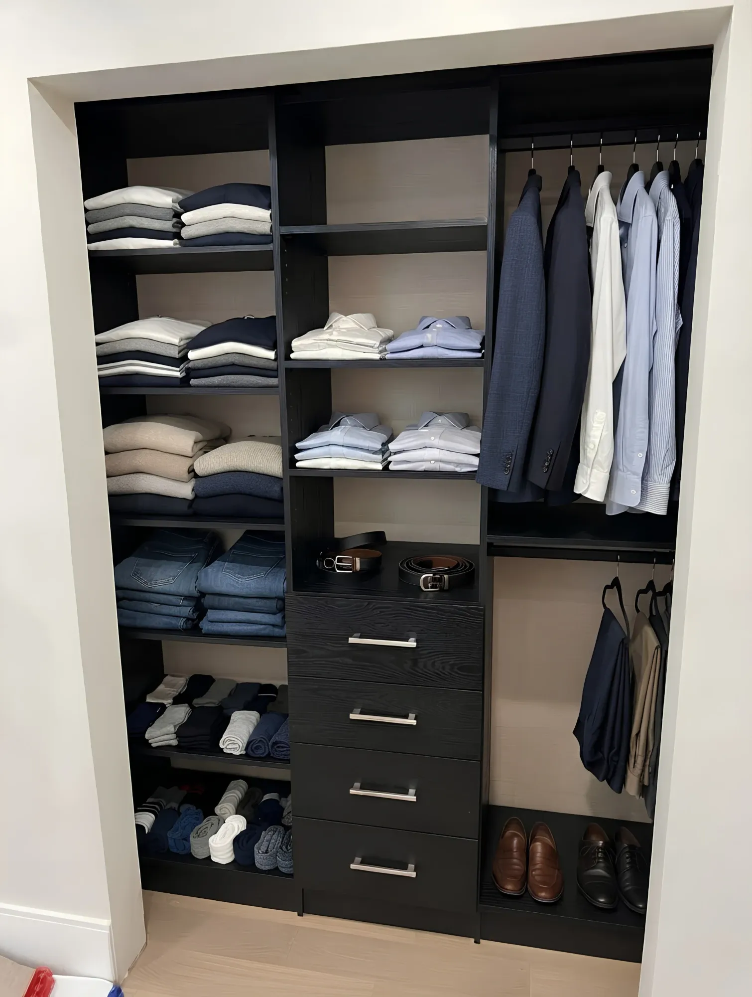 A black oak reach-in closet built into its opening, folded shelving on the left, shirts and jackets hung at two levels on the right and four drawers in the centre