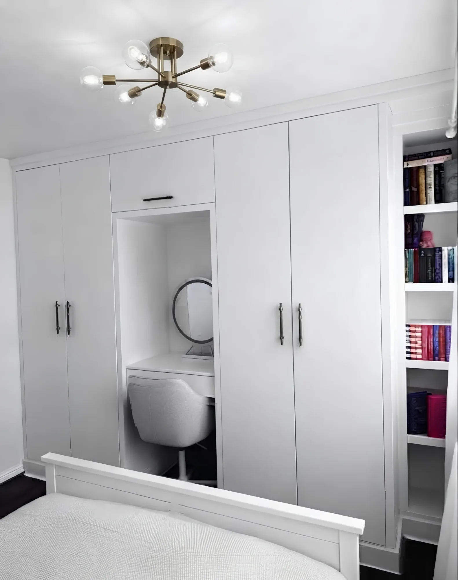 A white wardrobe wall built across a bedroom, full-height doors either side of a recessed vanity with a round mirror and a drawer under the counter