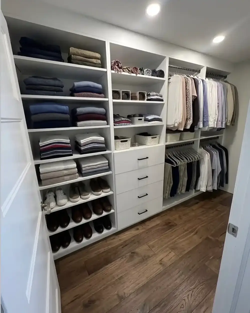 The corner of a custom white walk-in closet with shelves of folded sweaters and shoe shelving over a hardwood floor