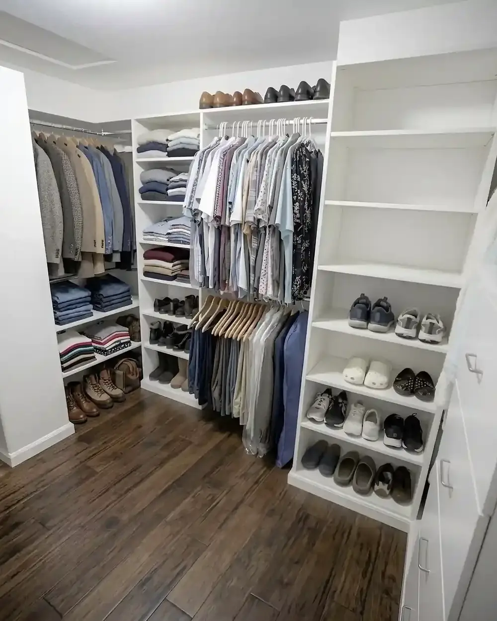 A white walk-in closet with long and short hanging sections and angled shoe shelves on a hardwood floor