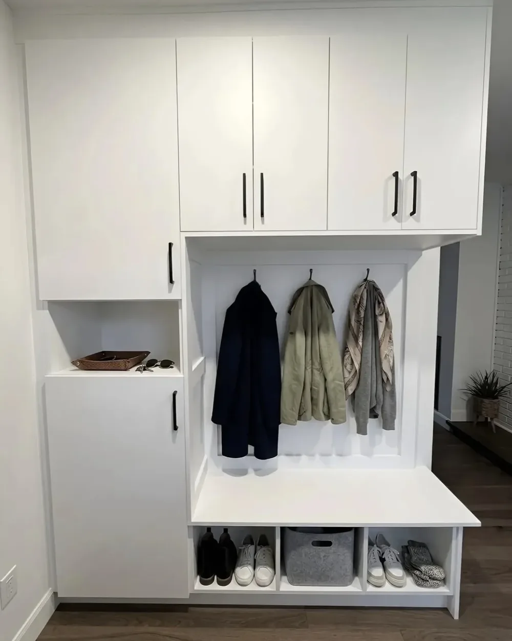 A white mudroom locker unit — a coat niche with three coats on hooks, closed cabinets above, a tall cupboard beside it and a bench over open cubbies of shoes