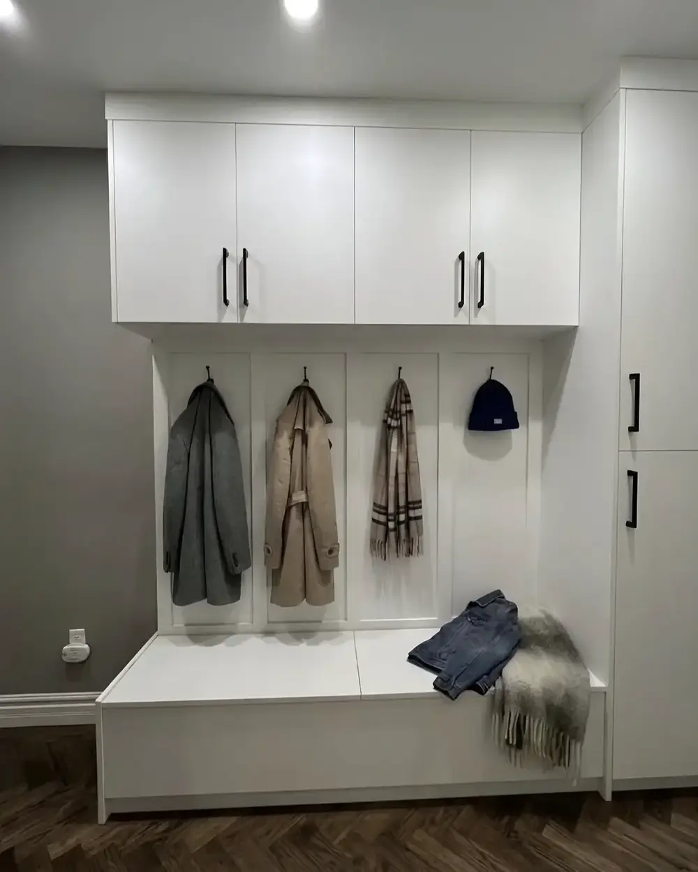 A compact white mudroom bench beside a garage door, with a cupboard above and coats on hooks