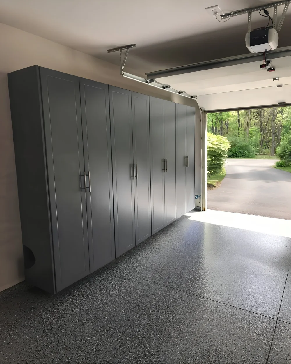 A full-height run of grey garage cabinets down one wall of a garage, floor to ceiling, on a speckled epoxy floor, with the door open to a driveway and trees beyond