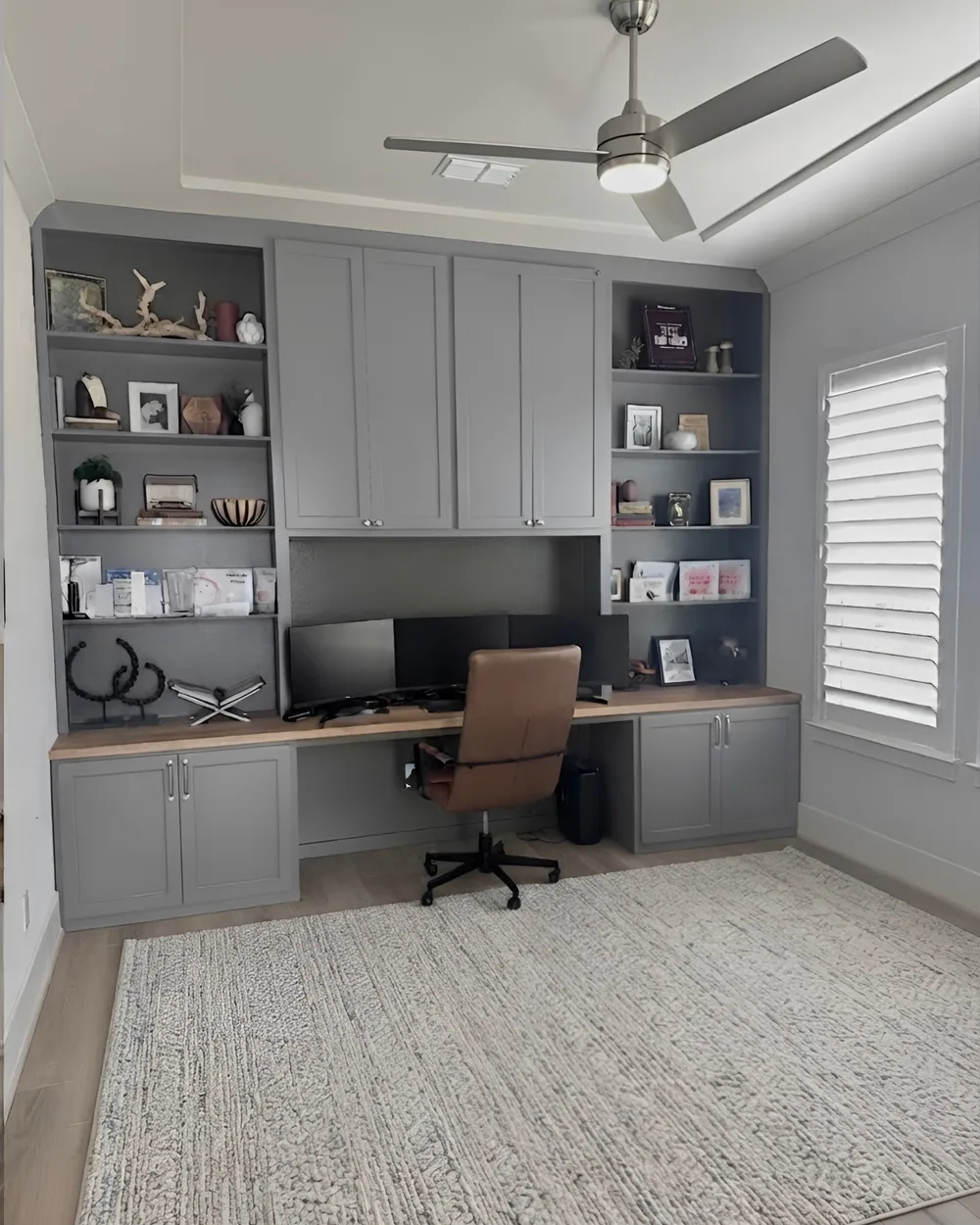 A grey built-in home office wall — a wood desktop running the width of the room between two lit bookcases, cabinets above and below, and a leather chair at the monitors