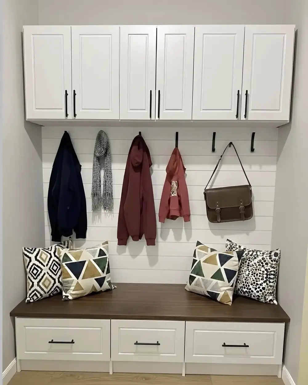 A white mudroom bench with cushions, coats on hooks and closed cabinets above