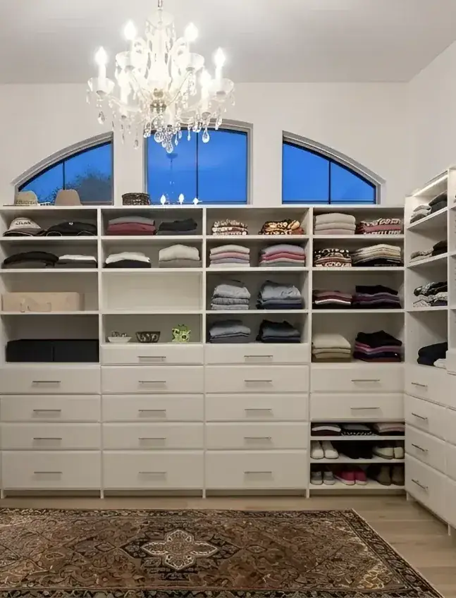 A wide custom walk-in closet in a Toronto home, hanging and shoe walls either side of a bank of drawers under an arched window