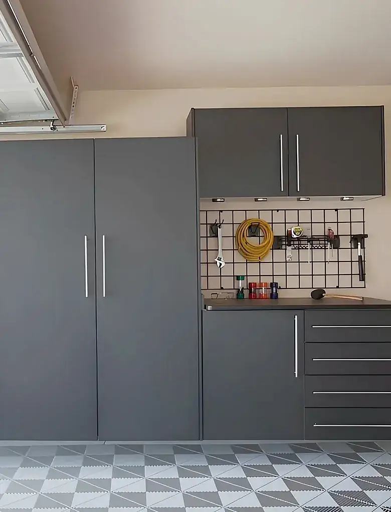 A double garage fitted with grey cabinets along the back wall — tall full-height units, a drawer bank under a worktop, and a wall-mounted tool panel hung with a hose and hand tools